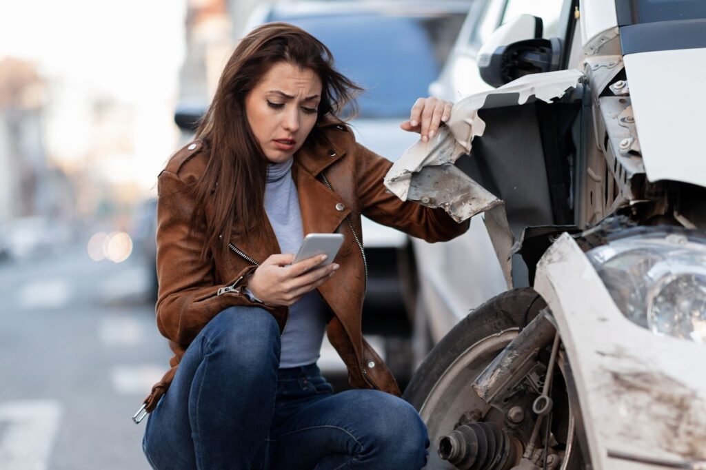 Young sad woman text messaging on phone after a car crash on the road